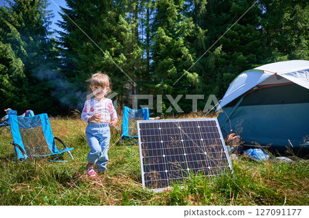 Young child playing near tourist tent with photovoltaic solar panel in summer. On background of dense forest. Integration of renewable energy in outdoor camping activities. Young child playing near tourist tent with photovoltaic solar panel in summer. On background of dense forest. Integration of renewable energy in outdoor camping activities. 127091177