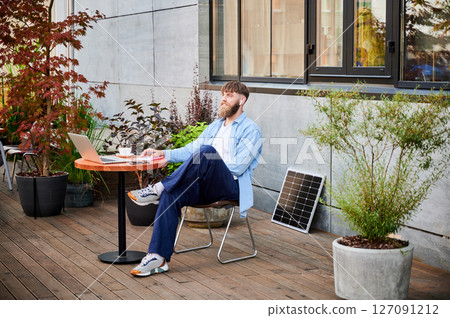 Happy bearded man works at round wooden table with laptop, notebook, and cup of coffee. Guy wears earbuds, surrounded by greenery and solar panel, highlighting modern, productive workspace. Happy bearded man works at round wooden table with laptop, notebook, and cup of coffee. Guy wears earbuds, surrounded by greenery and solar panel, highlighting modern, productive workspace. 127091212