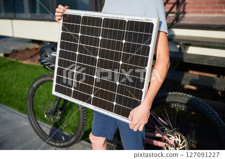 Close up view of person holding solar panel for charging, stands in front of brick building. Next to him pink electric mountain bike with black helmet resting on handlebars. Cropped view. 127091227