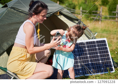 Young child and mother play with smartphone while charging with photovoltaic solar panel near tourist tent in summer. Integration of renewable energy in outdoor camping activities. Young child and mother play with smartphone while charging with photovoltaic solar panel near tourist tent in summer. Integration of renewable energy in outdoor camping activities. 127091228