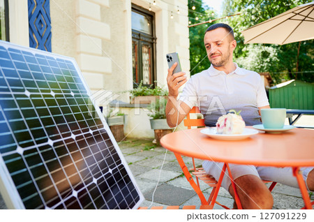 Young man charging smartphone from solar panel while sitting in cafe. Handsome male drinking coffee and checking his phone next to solar battery. Conscious male choosing sustainable lifestyle. 127091229
