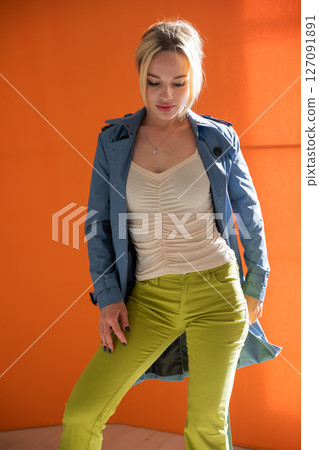 Portrait of a beautiful fashionable blonde woman in a blue raincoat on an orange background 127091891