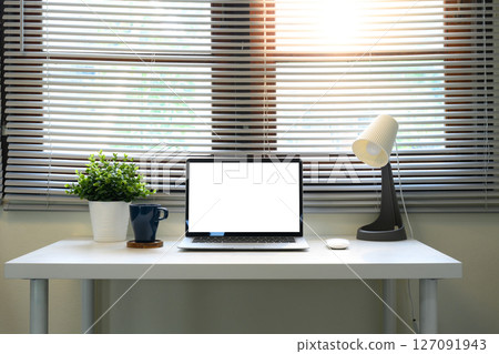 Bright home office with a laptop, coffee cup, indoor plants, and lamp under window blinds 127091943