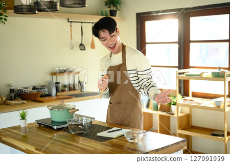 Happy man singing into a spoon while cooking in a bright kitchen 127091959