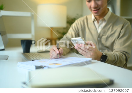 Smiling man checking his smartphone while writing notes on financial documents Smiling man checking his smartphone while writing notes on financial documents 127092007