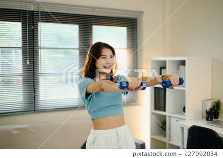 Cheerful young woman exercises with small dumbbells in her living room 127092034