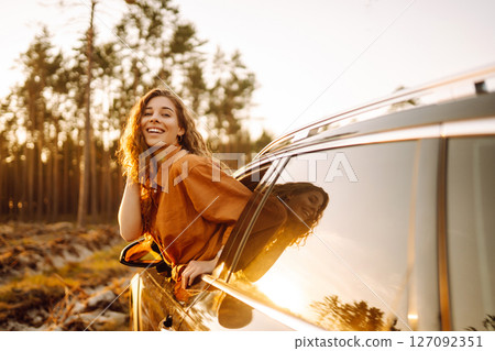 A young woman feels freedom leaning out of a car window in a sunny forest. Travel concept. A young woman feels freedom leaning out of a car window in a sunny forest. Travel concept. 127092351