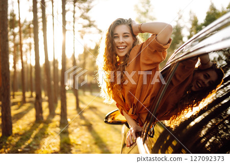 A young woman feels freedom leaning out of a car window in a sunny forest. Travel concept. 127092373