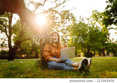 Portrait of a happy woman in sunny park on green lawn with a laptop. Freelance concept, technology. 127092456
