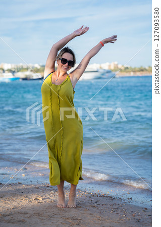 Woman in yellow dress stands against blue sea and beach waving her hands Woman in yellow dress stands against blue sea and beach waving her hands 127093580