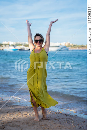 Woman in yellow dress standing against blue sea and beach with arms raised up Woman in yellow dress standing against blue sea and beach with arms raised up 127093584