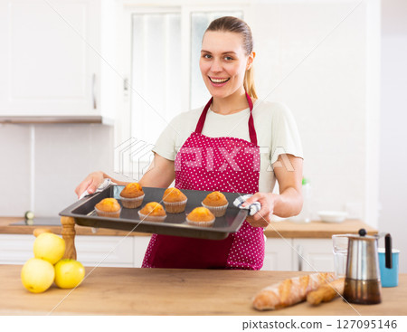 Smiling woman in apron standing in home kitchen with tray of cupcakes 127095146