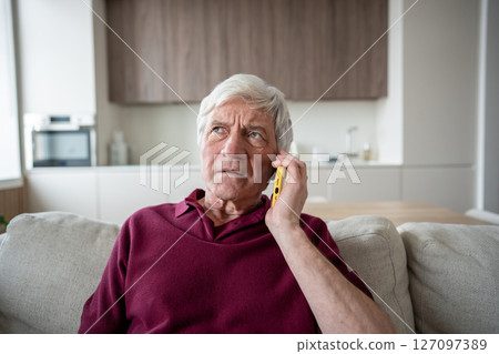 Suspicious anxious elderly man with phone at home, targeted by scam artists, telemarketing fake news 127097389