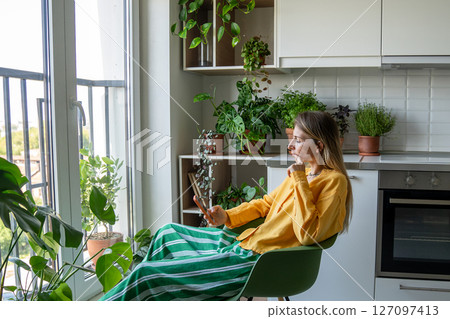 Thoughtful serious woman scroll news and social feeds on phone surrounded by indoor plants at home 127097413