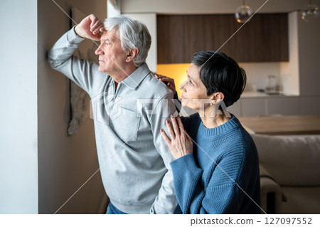 Thoughtful sad senior man look out window while wife reach out, consoling. Family support, empathy 127097552