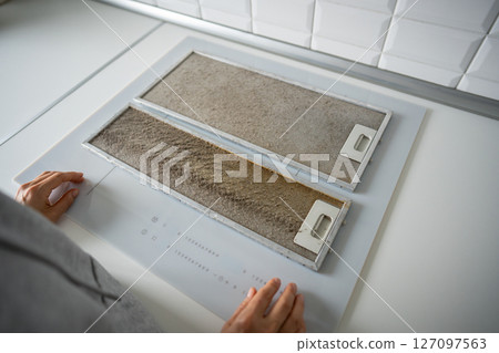 Greasy metallic kitchen hood filter needing deep cleaning. Homeowner hands near filter grid on table 127097563