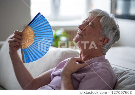 Heat can be deadly for seniors. Sweltering pensioner with fan struggling with summer heat indoors 127097608