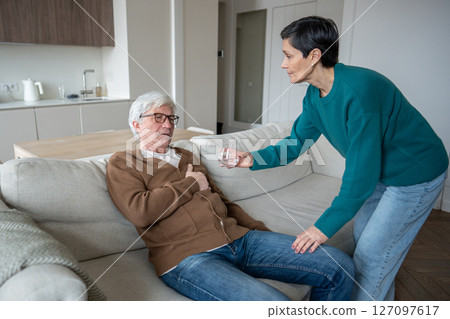 Caring wife helps elderly suffering husband clutching chest sitting on sofa, offering glass of water Caring wife helps elderly suffering husband clutching chest sitting on sofa, offering glass of water 127097617
