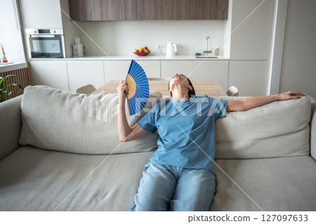 No cooling system, elderly woman struggling without air conditioning, resting on couch using fan 127097633