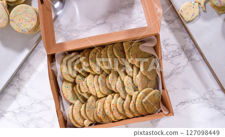 Flat lay. With precision, the woman is carefully arranging the sugar cookies, filled with dough-mixed sprinkles, into a rustic brown paper box. 127098449
