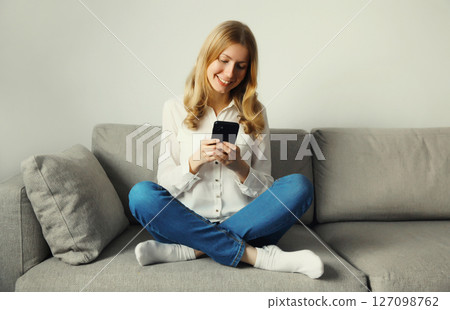 Happy young woman looks at phone, girl with smartphone sitting on the couch in room at home Happy young woman looks at phone, girl with smartphone sitting on the couch in room at home 127098762