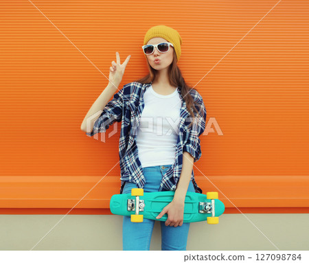 Portrait of stylish young girl with skateboard in hat, happy modern teenage female posing on orange Portrait of stylish young girl with skateboard in hat, happy modern teenage female posing on orange 127098784