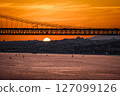Beautiful sunset over 25 de Abril bridge in silhouette and Tagus river with sailboats drifting below and Lisbon skyline in summer haze in the background. Orange sky, clouds and sun, tranquil waters 127099126