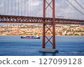Colorful landscape with blue container ship on Tagus river under iconic 25 de Abril or 25th April bridge, Lisbon, Portugal. Famous ironwork of red suspension bridge, vibrant cityscape, urban 127099128