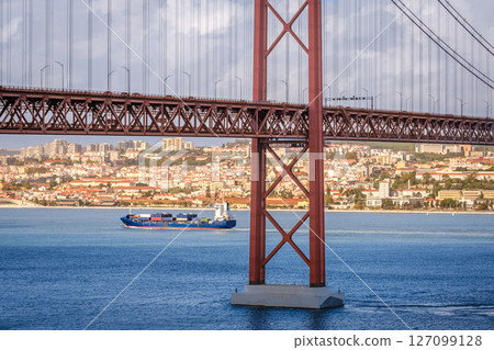 Colorful landscape with blue container ship on Tagus river under iconic 25 de Abril or 25th April bridge, Lisbon, Portugal. Famous ironwork of red suspension bridge, vibrant cityscape, urban 127099128