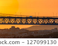 Beautiful view of golden sunset and suspension city bridge with its steel structure and cables, dramatic silhouettes of vehicles in motion blur against bright orange sky. Distant hills and urban 127099130