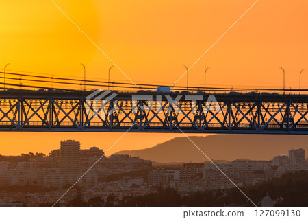 Beautiful view of golden sunset and suspension city bridge with its steel structure and cables, dramatic silhouettes of vehicles in motion blur against bright orange sky. Distant hills and urban 127099130