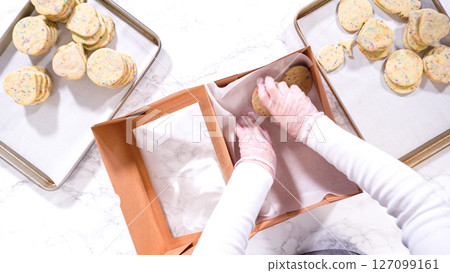 Flat lay. With precision, the woman is carefully arranging the sugar cookies, filled with dough-mixed sprinkles, into a rustic brown paper box. Flat lay. With precision, the woman is carefully arranging the sugar cookies, filled with dough-mixed sprinkles, into a rustic brown paper box. 127099161