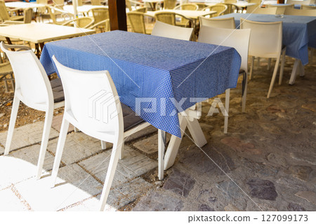 Empty wicker chairs in outdoor cafe under trees. Hospitality service, seasonal tourism, and summer leisure in public spaces. 127099173
