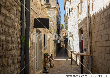 Stone alleyway with decorative figures and empty cafe tables in Mediterranean old town. Local charm, tourism atmosphere, and European summer. Stone alleyway with decorative figures and empty cafe tables in Mediterranean old town. Local charm, tourism atmosphere, and European summer. 127099181