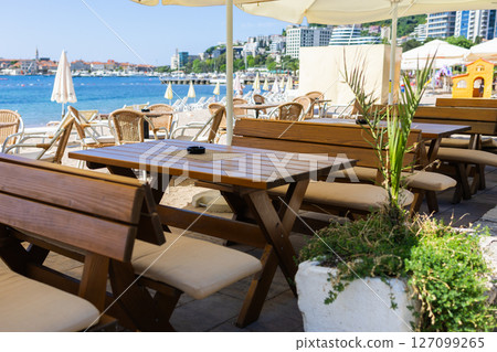 Wooden benches and tables at beach cafe under umbrella. Coastal hospitality, summer relaxation, and outdoor seating near sea. Wooden benches and tables at beach cafe under umbrella. Coastal hospitality, summer relaxation, and outdoor seating near sea. 127099265