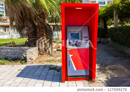 Red ATM machine stands outdoors near palm tree and walkway. Urban banking, financial access, and everyday public service infrastructure. Red ATM machine stands outdoors near palm tree and walkway. Urban banking, financial access, and everyday public service infrastructure. 127099278
