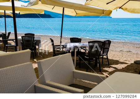 Tables with chairs under umbrellas on sandy beach. Summer tourism, seaside dining, and vacation leisure in coastal destination Tables with chairs under umbrellas on sandy beach. Summer tourism, seaside dining, and vacation leisure in coastal destination 127099284