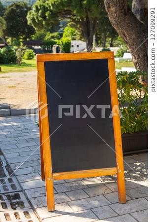 Wooden frame chalkboard stands on summer pavement. The concept shows outdoor signage, seasonal business, and blank advertising opportunity. Copy space. Wooden frame chalkboard stands on summer pavement. The concept shows outdoor signage, seasonal business, and blank advertising opportunity. Copy space. 127099291