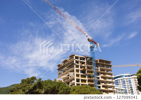 Construction crane above residential building under blue sky. Urban infrastructure, civil engineering, and real estate development Construction crane above residential building under blue sky. Urban infrastructure, civil engineering, and real estate development 127099341