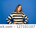 Pretty Hispanic teenage girl wearing a black and white striped sweater with her arms akimbo, waiting for something, isolated on a plain blue studio background. Lifestyle concept. 127101107