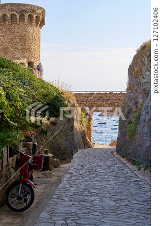 A scenic pathway in Tossa de Mar leads towards the sea, with a red moped parked alongside the stone walls. The stone tower adds historical charm. 127102406