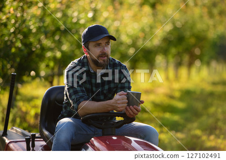 Farmer sitting on small tractor in orchard, enjoying peaceful moment. 127102491