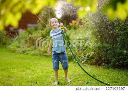 Funny preteen boy pouring cool water on himself and playing with garden hose with sprinkler in sunny backyard on hot summer day. Happy child having fun with water splashes. 127102697