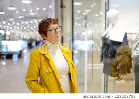 Portrait of elderly woman during shopping at mall. Beautiful red-haired senior lady chooses clothes for herself while looking at new models on mannequins in a showcase 127102792
