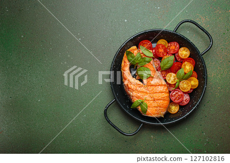 Grilled salmon steak with cherry tomatoes and basil leaves in black pan with cutlery top view on dark green background, space for text 127102816