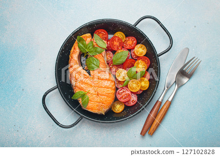 Grilled salmon steak with cherry tomatoes and basil leaves in black pan with cutlery top view on light blue background Grilled salmon steak with cherry tomatoes and basil leaves in black pan with cutlery top view on light blue background 127102828