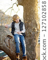 A cute preteen boy is sitting on a branch of a large sycamore tree 127102858