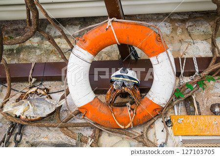 Weathered lifebuoy with humorous crab in sailor hat on nautical-themed wall. Coastal decor, marine symbolism, and playful maritime detail. Weathered lifebuoy with humorous crab in sailor hat on nautical-themed wall. Coastal decor, marine symbolism, and playful maritime detail. 127103705