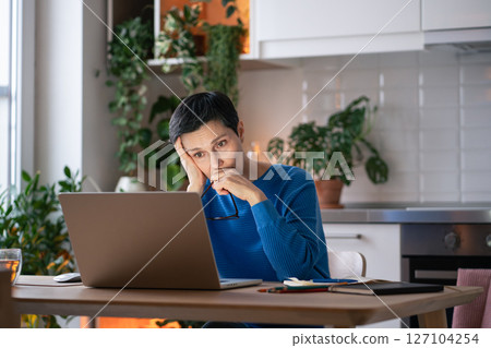 Exhausted burdened mature woman looks at laptop screen at home, with notebook of notes nearby 127104254