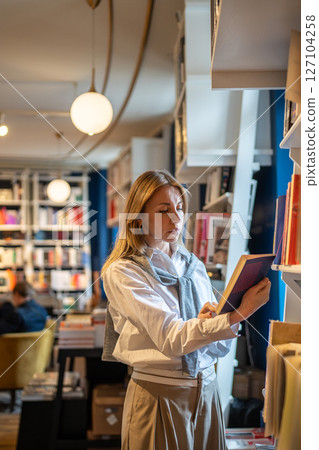 Focused middle aged stylish woman choosing book in bookstore, standing near window reads content. Focused middle aged stylish woman choosing book in bookstore, standing near window reads content. 127104258